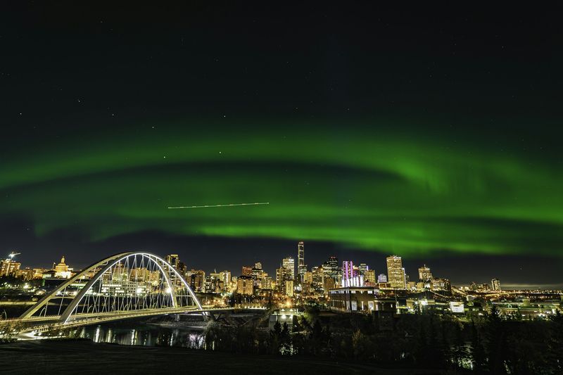 Green northern lights over the downtown Edmonton skyline and the white arches of the Walterdale Bridge at night.